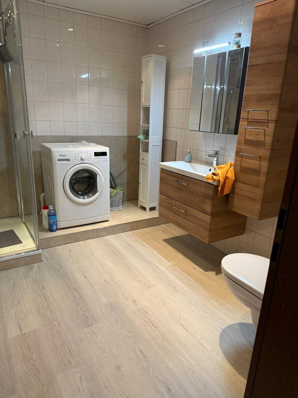 Badezimmer mit Waschtisch und Hochschrank in Holzoptik, beleuchtetem Spiegelschrank und Duschkabine