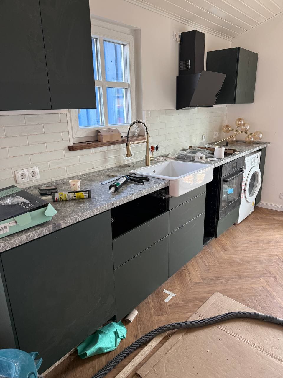 Dunkelgrüne Küche während der Montage mit weißer Keramikspüle, goldfarbener Armatur, Arbeitsplatte in Steinoptik und schräger Dunstabzugshaube