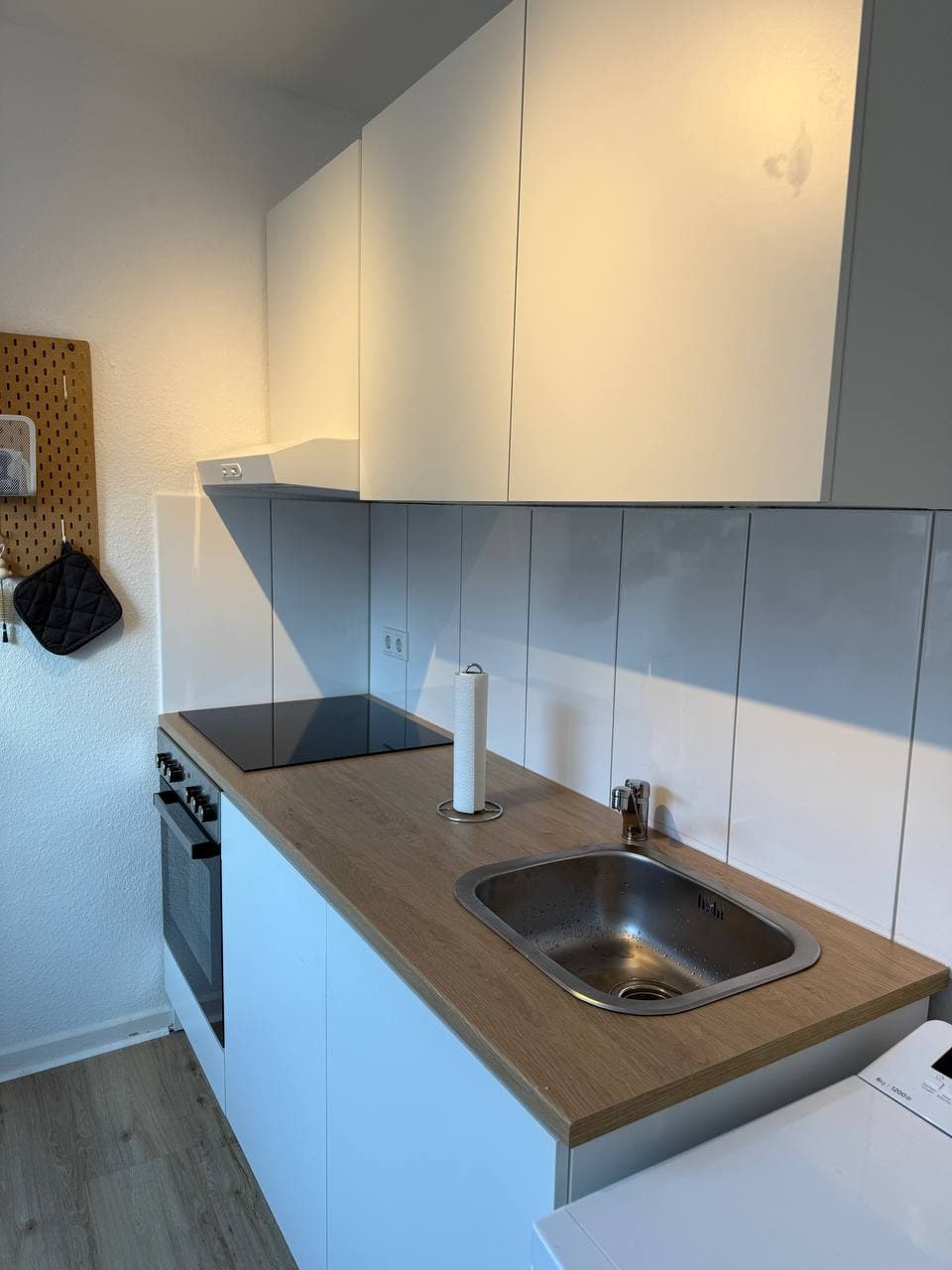 Kleine weiße Küche mit grifflosen Fronten, Arbeitsplatte in Holzoptik, Edelstahlspüle, Kochfeld und Dunstabzugshaube